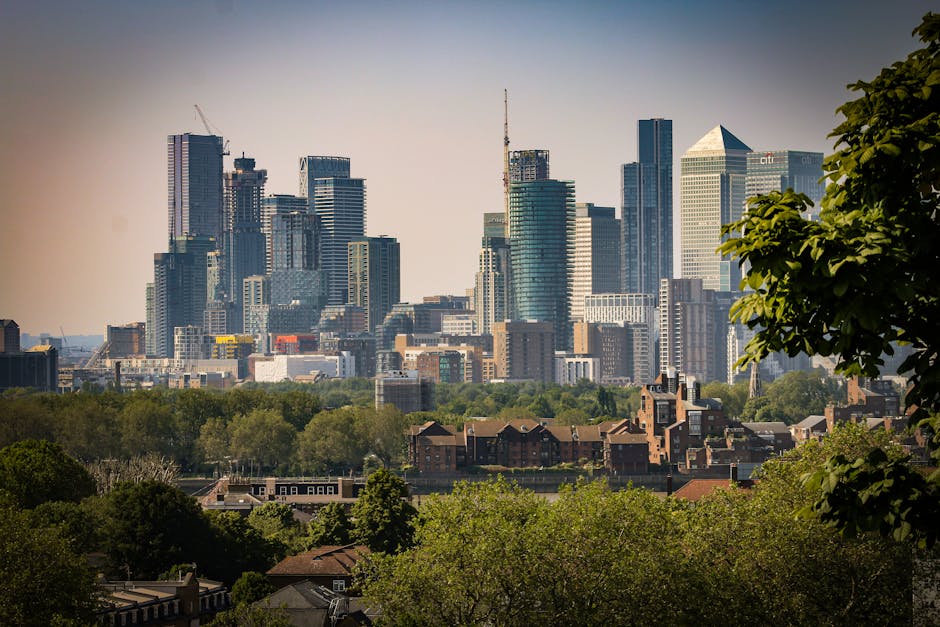 The image shows a city skyline with numerous modern skyscrapers, including high-rise office buildings and residential towers, with a mix of glass and steel facades reflecting the daylight. In the foreground, there is a residential area with red-brick apartment buildings and houses surrounded by lush green trees, which provide a natural contrast to the urban environment. The buildings in the distance vary in height and architectural style, some still under construction with cranes visible atop them. The sky above is clear with a subtle gradient from light to darker blue, suggesting a calm, sunny day. The scene is taken from a vantage point overlooking a park or green space area at the lower part of the image, indicating a separation between the city's greenery and dense commercial development, relevant for visualising urban expansion and the contextual environment in which private or independent waste removal services like those offered by Rubbish Clearance Greenwich might operate within city boundaries.