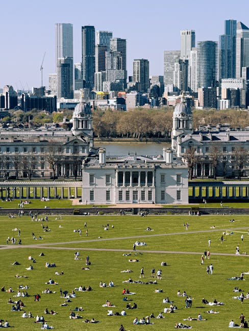 A historic stone building with an ornate domed tower featuring a golden weather vane at the top is located in the foreground of the image, with a black wrought-iron fence running along its perimeter. The building has classical architectural elements, including columns and decorative stonework, and is set on a neatly maintained grassy area with a few benches and pedestrians visible nearby. In the background, there is a modern city skyline composed of high-rise office buildings and skyscrapers made of glass and steel, reflecting sunlight against a partly cloudy sky with scattered white clouds. The scene captures a contrast between the older architectural style of the foreground structure and the contemporary urban skyline behind it, possibly illustrating an area where private or independent waste clearance services operate in a city setting, supporting alternative waste handling arrangements outside of council-run rubbish collection.
