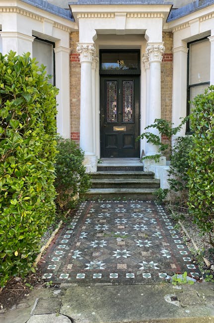 The image depicts the entrance to a traditional brick-built house with classical architectural features, including white decorative columns flanking the black front door, which has stained glass panels and a brass mail slot. In front of the door, there is a small set of weathered stone steps leading up from a patterned tiled pathway composed of dark and light stones forming geometric designs. Lush green bushes border the pathway on both sides, with the left bush appearing more prominent, and the right bush extending slightly towards the entrance. The exterior walls of the house are constructed from yellowish-brown bricks, with white trim accents around the windows and roofline. The scene is well-lit, suggesting daylight, and the overall setting appears neat and well-maintained, indicative of a private residence. Although the image does not explicitly show rubbish or waste, the tidy and organized appearance aligns with professional waste management or clearance services, such as those provided by Rubbish Clearance Greenwich in the context of house clearances or property maintenance.