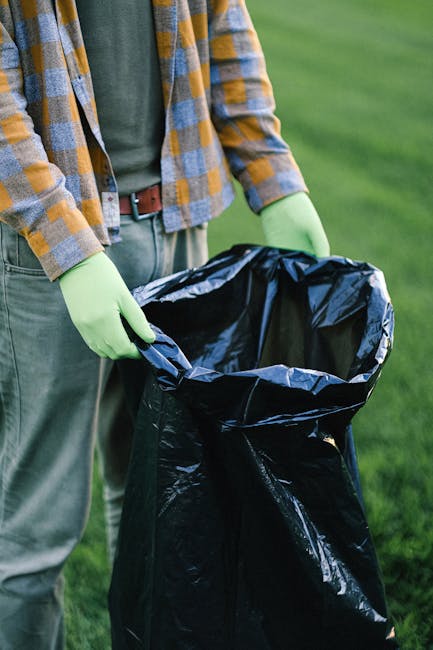 A person wearing a yellow and grey checkered shirt over a green top and grey trousers, with bright green gloves, is holding open a large black rubbish bag lined with a silver inner layer. The person appears to be in an outdoor setting on a grassy area, possibly a garden or park. Visible only from the waist up, the individual is standing next to a neatly maintained lawn, with no other objects or background details visible. The black plastic bag is partially filled, with its opening held open for rubbish collection or waste disposal activities, which aligns with private waste handling services. The scene suggests an activity related to rubbish removal or garden waste clearance, occurring in a setting suitable for on-site waste collection or environmental maintenance, which is relevant to independent or professional rubbish clearance services.