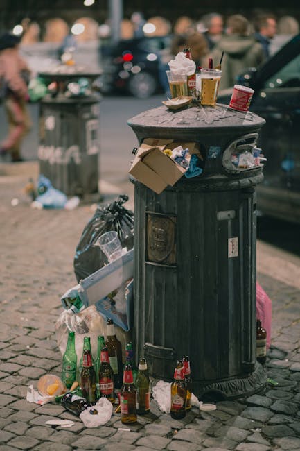 A collection of overflowing rubbish and waste materials situated on the pavement beside a parking area, featuring various discarded items including black and black plastic bin bags, open cardboard boxes with packing materials, paper and plastic packaging, and loose paper strewn across the ground. Behind the waste, there are three wheelie bins—one grey open container filled with mixed paper and cardboard, a black bin, and a red bin—positioned near a metal railing. To the left, a parked silver hatchback car is partially visible, with a yellow license plate. In the background, a building with a multi-storey facade is under construction or renovation, covered in blue scaffolding and protective sheeting, with storefronts on the ground level, including a shop with a bright yellow sign. The environmental context suggests an urban area where private or independent rubbish clearance might be necessary, supported by waste management services such as Rubbish Clearance Greenwich, who handle diverse rubbish types for streets and estates in the SE10 vicinity. The overall scene emphasizes the importance of waste disposal and rubbish removal to maintain cleanliness in public spaces.