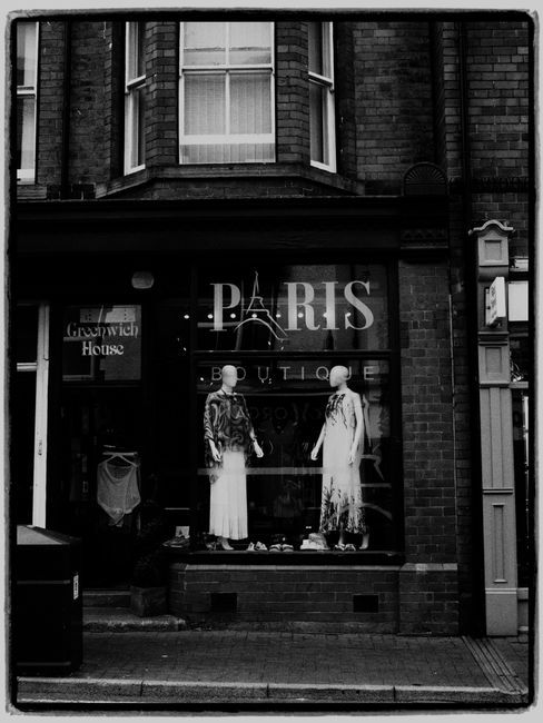A black and white photograph of a boutique shopfront situated within a brick building, capturing the display window and entrance area. The large glass window features the partially visible text 'PARIS' in bold, white lettering, with additional smaller words beneath that are obscured. Inside the window, there are two mannequins dressed in long, flowing garments; one wearing a patterned blouse with wide-leg trousers, and the other in a lighter, more delicate outfit, positioned on either side of the display. To the left of the mannequins, a clothing rack holds garments, including a prominent dress or top hanging on a hanger. The street scene shows a well-maintained sidewalk with a small black waste bin partially visible on the left, and the shop's brick exterior with a dark, textured finish. The building features a curved bay window on the upper level, with double-pane sash windows and white curtains visible inside. The overall atmosphere is subdued, with natural lighting creating soft contrasts that highlight the textures of the brickwork, glass, and clothing displays, subtly reflecting the idea of private or alternative waste handling through the unobstructed shopfront setting associated with urban rubbish removal services.