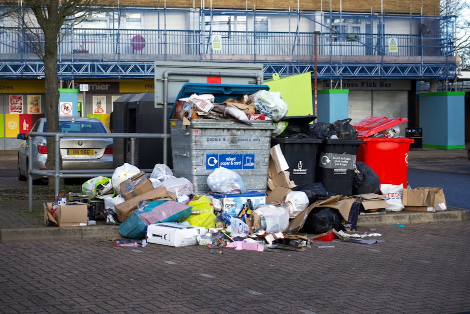 A large pile of mixed waste and rubbish bags, consisting of cardboard boxes, paper, plastic bags, and various discarded packaging materials, situated on a paved sidewalk near a metal railing. The trash is overflowing from multiple bin containers, including a grey bin labeled for mixed paper and card, a black bin for general waste, a red bin, and a green recycling bin, which are all positioned close together. Surrounding the rubbish pile, there are parked cars, a tree with a bare trunk on the left side, and a background of a commercial building with blue scaffolding and signage. Visible litter spills onto the pavement, highlighting the accumulation of waste outside a commercial property. This scene emphasizes the importance of proper rubbish removal and waste management, which services such as Rubbish Clearance Greenwich can provide for local businesses seeking alternative or private waste disposal solutions.