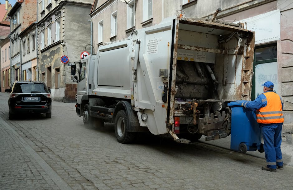A street scene featuring a white rubbish collection truck with an open rear loading compartment, which appears to be in the process of collecting waste. The truck is parked on a cobblestone pavement adjacent to a row of older, multi-story buildings with varying architectural details and weathered facades. A worker dressed in a blue uniform and high-visibility orange vest, also wearing a blue cap and gloves, is positioned on the right, operating a blue wheelie bin and preparing to empty it into the truck’s compartment. In the background, a black car is parked closer to the curb, and there are two traffic signs mounted on a pole on the building facade, indicating parking restrictions. The scene is illuminated by natural daylight, with a neutral, urban environment that reflects typical waste collection activity, supporting the context of alternative rubbish removal methods outside of council-organized disposal procedures.