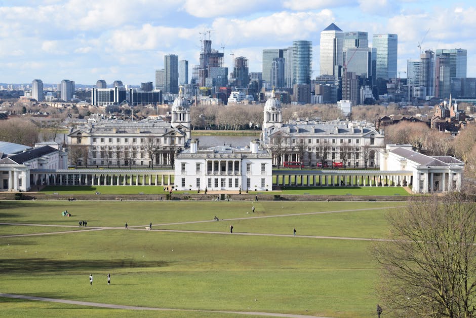 A wide view of a historic white neoclassical building with symmetrical towers and colonnaded wings, situated in a large landscaped park with expansive green lawns and walking paths, with small groups of people scattered across the grass. In the background, a modern city skyline featuring tall glass skyscrapers and construction cranes is visible under a partly cloudy sky, contrasting with the classical architecture in the foreground. The scene is captured during daytime with natural lighting emphasizing the details of the building's façade and the lush greenery surrounding it. The image, related to waste management services, subtly highlights the contrast between historic and contemporary urban environments, relevant for clients interested in alternative waste removal or private disposal options featuring on-site clearance or independent collection methods, such as those provided by Rubbish Clearance Greenwich.