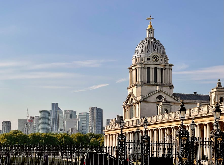 A detailed view of a historic classical-style building featuring a large central dome topped with a golden weather vane. The building's facade showcases columns and ornate architectural details, with a black wrought-iron fence in the foreground. To the right and behind the main structure, additional architectural elements are visible. In the background, a modern city skyline with tall glass skyscrapers contrasts with the older architectural style of the foreground building. The scene is set in daylight with a clear blue sky and some wispy clouds, highlighting the textures of the stonework and the metallic sheen of the dome. The area appears well-maintained with greenery at the base of the building, which is typical of historic or civic structures often located in urban environments—an appropriate setting for visual content related to independent or private waste collection and rubbish removal services, as it emphasizes the blend of historical architecture and contemporary cityscape.