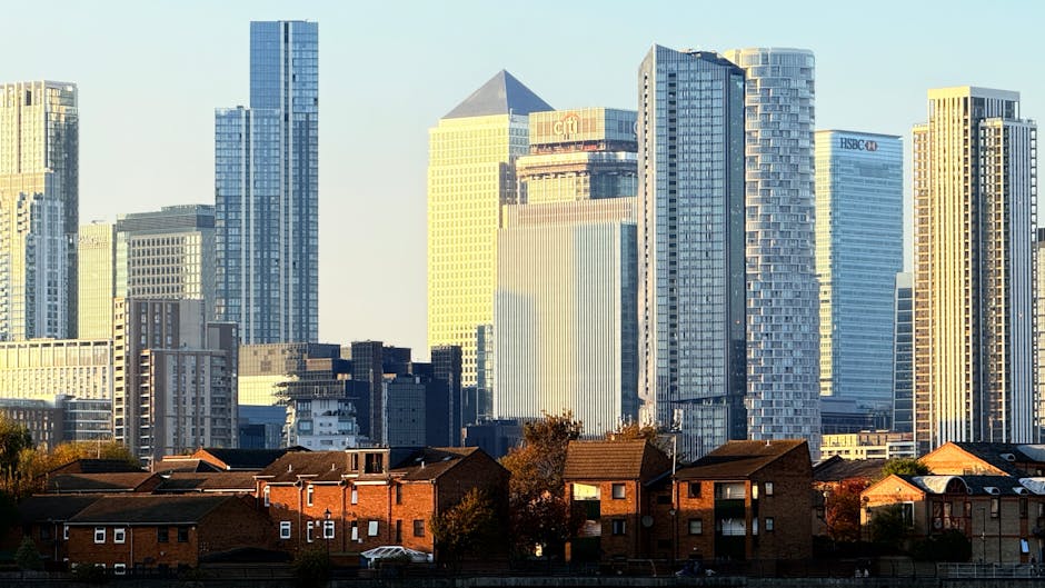 The image depicts a panoramic view of a modern city skyline with numerous high-rise buildings and skyscrapers, some still under construction with visible cranes atop them. The buildings vary in height, shape, and glass facades, reflecting sunlight, with a mix of rectangular and curved architectural designs. In the foreground, there is a lush, green park area featuring numerous mature trees with dense foliage, providing a natural contrast to the urban environment. The trees display different shades of green, and some branches extend into the frame, overlapping part of the cityscape. The sky overhead is mostly clear with a few wispy clouds, contributing to bright, natural daylight that illuminates both the city and the greenery. The scene suggests a peaceful, outdoor setting near a densely populated business district, aligning with the theme of private waste handling and urban rubbish management, as might be associated with independent rubbish removal services in a city context, with the natural environment providing a buffer zone around the urban core. The overall composition highlights the contrast between natural and constructed elements, indicative of a typical city park overlooked by modern commercial buildings.