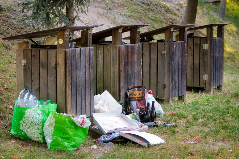 A row of four wooden outdoor rubbish bins with slanted roofs, positioned on a grassy area adjacent to a wooded hillside. The bins are made of weathered, dark-stained vertical wooden planks, each secured with metal hinges and latches. Trash, including a green plastic bag filled with empty plastic bottles and other waste items, is spilled onto the ground in front of the bins. There is a white pizza box, several glass bottles, plastic bags, and miscellaneous litter scattered on the grass. The background shows a gently sloping hillside with trees and greenery, suggesting a private outdoor space or park area where waste has accumulated outside the designated bins. The scene is illuminated by natural daylight, emphasizing the contrast between the neatly constructed bins and the scattered rubbish that appears to be awaiting collection, indicative of an on-site waste management setup often used for independent rubbish removal services such as those provided by Rubbish Clearance Greenwich.