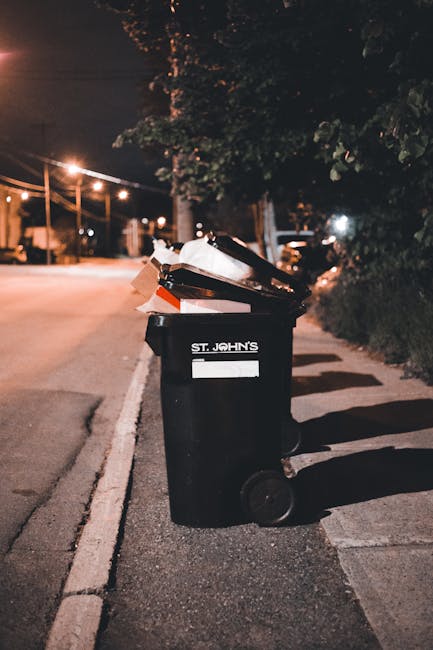 A row of four wooden outdoor rubbish bins with slanted roofs, positioned on a grassy area adjacent to a wooded hillside. The bins are made of weathered, dark-stained vertical wooden planks, each secured with metal hinges and latches. Trash, including a green plastic bag filled with empty plastic bottles and other waste items, is spilled onto the ground in front of the bins. There is a white pizza box, several glass bottles, plastic bags, and miscellaneous litter scattered on the grass. The background shows a gently sloping hillside with trees and greenery, suggesting a private outdoor space or park area where waste has accumulated outside the designated bins. The scene is illuminated by natural daylight, emphasizing the contrast between the neatly constructed bins and the scattered rubbish that appears to be awaiting collection, indicative of an on-site waste management setup often used for independent rubbish removal services such as those provided by Rubbish Clearance Greenwich.