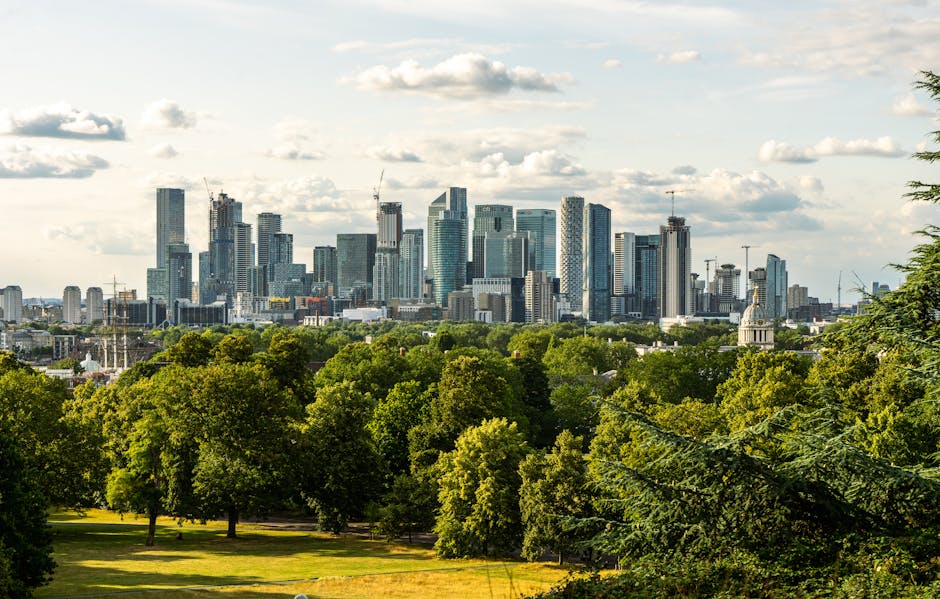 A wide view of a modern city skyline with numerous tall office buildings and skyscrapers made of glass and steel, featuring a variety of reflective surfaces and contemporary architectural styles, situated behind a large green park area with lush, leafy trees and open grassy spaces in the foreground. The sky above is partly cloudy with patches of sunlight illuminating the buildings and the park, creating a balanced natural and urban scene. The image highlights the contrast between the natural environment of the park and the dense, high-rise development of the city, typical of an alternative waste handling approach where urban rubbish removal may be carried out independently of municipal services, with a focus on curbside and on-site clearance. Rubbish Clearance Greenwich can provide professional rubbish removal solutions in such urban environments, supporting sustainable and efficient waste management practices amid high-density cityscapes.