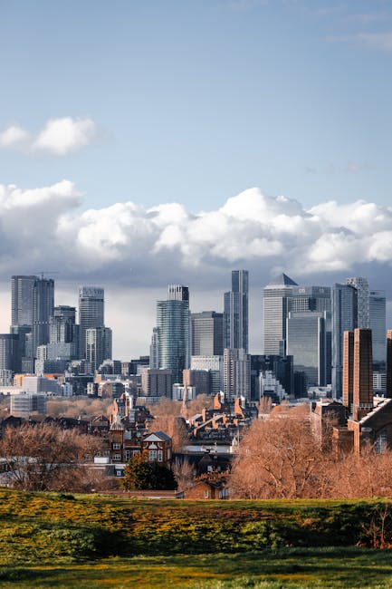 A wide view of a city skyline with numerous modern high-rise office buildings and skyscrapers made of glass and steel, set against a partly cloudy sky with large, white, fluffy clouds. In the foreground, there is a grassy area with a low hedge and some leafless trees, indicating a park or green space adjacent to urban development. The buildings vary in height and design, with some featuring reflective glass surfaces that capture the natural light. The scene is taken during daytime with clear weather, highlighting the contrast between the natural environment and the dense cluster of modern urban structures, reflecting typical scenes where private waste collection or on-site clearance services such as those provided by Rubbish Clearance Greenwich may operate near city centers.